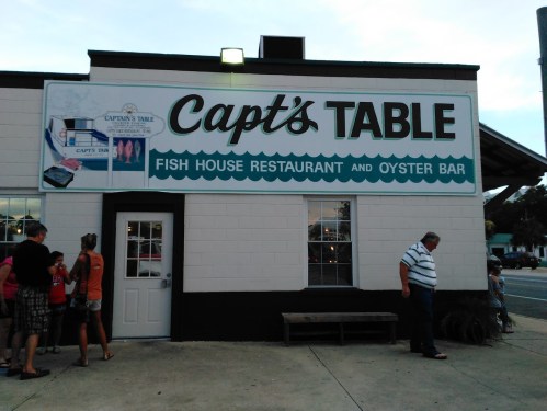 Sign for "Capt's Table Fish House Restaurant and Oyster Bar" on a building, with people gathered outside.