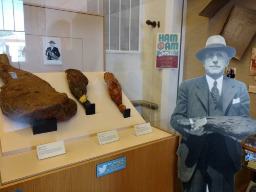 A display featuring large cured hams, a vintage photo, and a life-sized cutout of a man in a suit and hat.