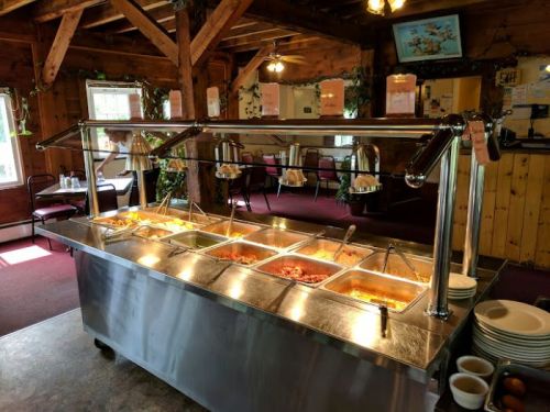 A buffet table with multiple food trays under glass covers in a rustic dining area with wooden beams.