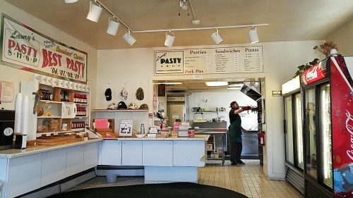 Interior of a pasty shop with a counter, menu board, and a worker preparing food in the kitchen area.