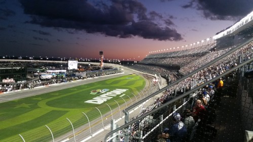 A panoramic view of a racetrack at dusk, filled with spectators and illuminated grandstands.