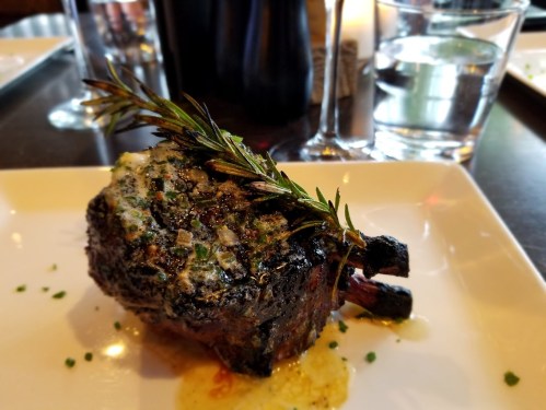 A beautifully plated lamb chop topped with herb butter and garnished with a sprig of rosemary on a white plate.