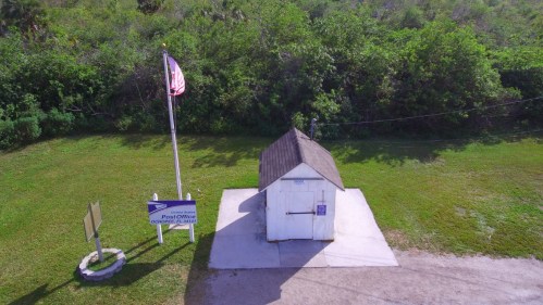 A small white post office building with a flag, surrounded by green grass and trees.