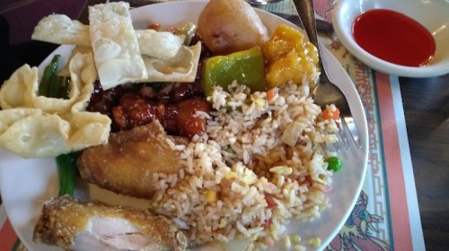 A plate of assorted Chinese food including fried rice, chicken, spring rolls, and sweet and sour sauce.
