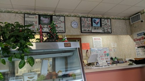 Interior of a restaurant with a menu board, clock, and decorative plants. A counter is visible in the foreground.