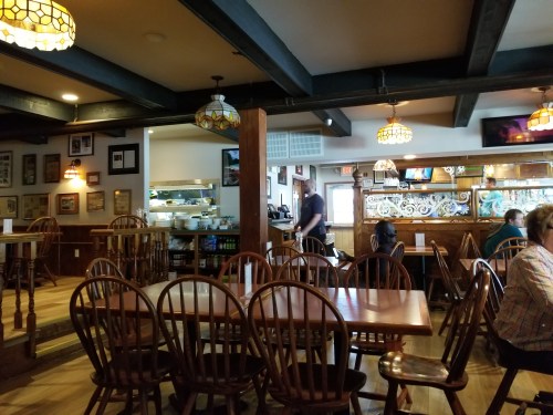 Interior of a cozy restaurant with wooden tables, chairs, and warm lighting. A kitchen area is visible in the background.