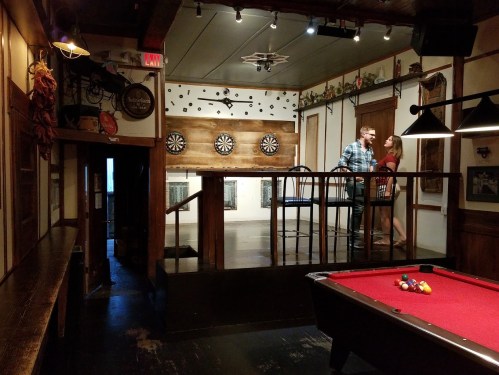 A cozy bar with dartboards and a pool table, featuring a couple chatting near the stage area.