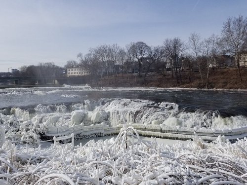 A frozen river with icy banks and a waterfall, surrounded by bare trees and a distant building under a clear sky.