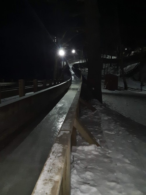 A snowy, illuminated sledding hill at night, with a winding path and wooden railings.