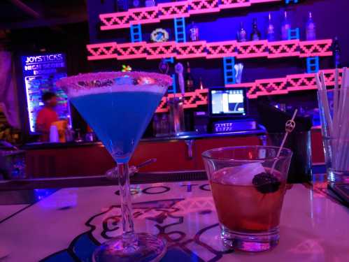 Two colorful cocktails on a bar: a blue martini with a sugared rim and a brown drink with a cherry and straw.