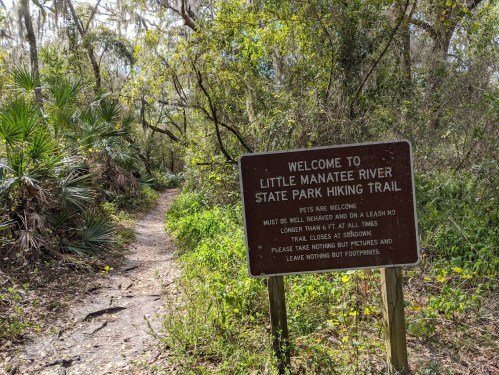 A sign at the entrance of Little Manatee River State Park hiking trail, surrounded by lush greenery and a dirt path.