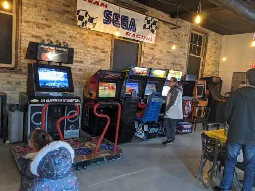 A retro arcade with several racing game machines and people playing, featuring a Sega Racing banner above.