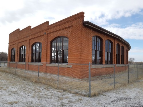 A red brick building with large windows, surrounded by a chain-link fence, set against a cloudy sky.