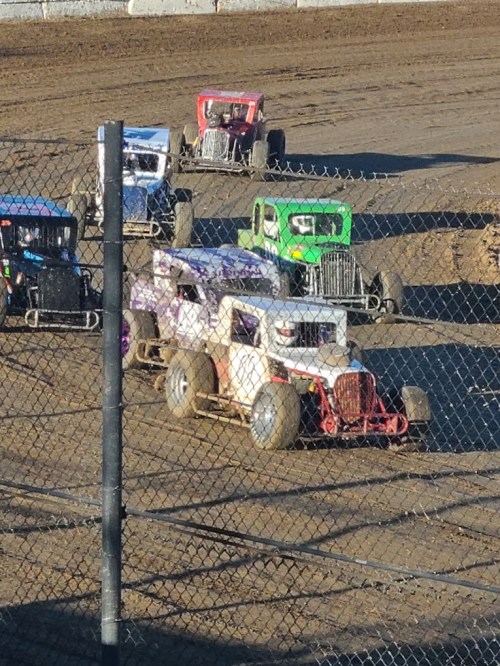 A group of colorful dirt track racing cars compete on a muddy track, with a fence in the foreground.