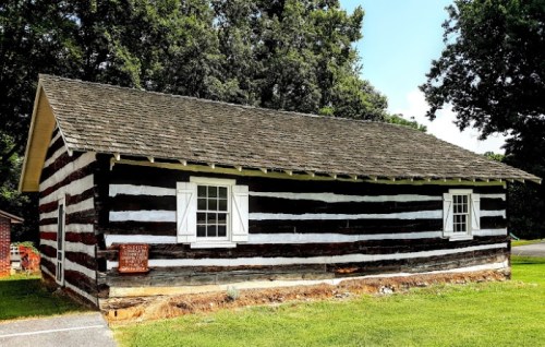 A rustic log cabin with a striped pattern, featuring white windows and a sloped roof, surrounded by greenery.