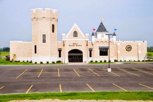 A large, castle-like building with towers and colorful flags, surrounded by a parking lot and green fields.