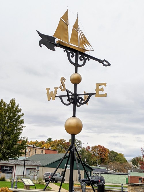 A decorative weather vane featuring a ship, with directional letters W, S, E, and a central gold sphere.
