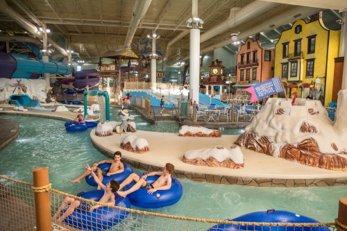 Indoor water park scene with people floating on tubes, water slides, and colorful buildings in the background.