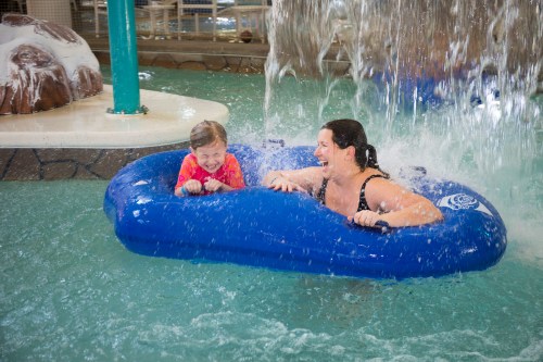 A woman and a child enjoy a splash in a blue inflatable raft under a waterfall in a pool. Both are smiling and having fun.