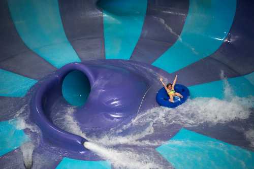 A child rides a blue inner tube down a colorful water slide, splashing through water with arms raised in excitement.