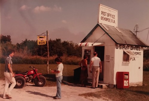 A small, vintage post office in Ochopee, Florida, with people and an ATV nearby, set in a rural landscape.
