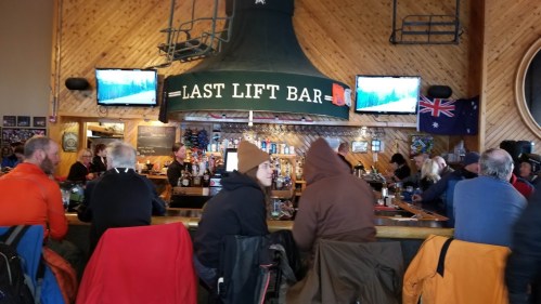 A cozy bar scene with patrons seated at the counter, watching screens, surrounded by wooden decor and ski-themed elements.