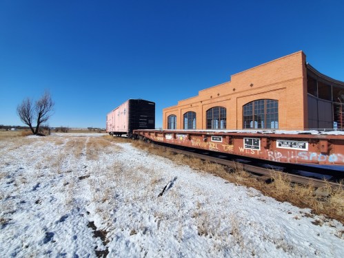 A pink train car next to a brick building, with a snowy field and clear blue sky in the background.