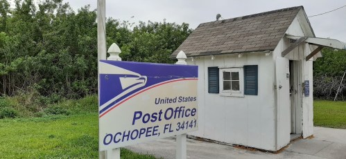 A small white building with a sign reading "United States Post Office, Ochopee, FL 34141" in a grassy area.