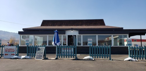 A restaurant with a blue umbrella and a fenced patio, featuring a sign and snow bags in front on a clear day.