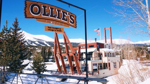 Sign for Ollie's Pub & Grub in a snowy landscape, with mountains and trees in the background.