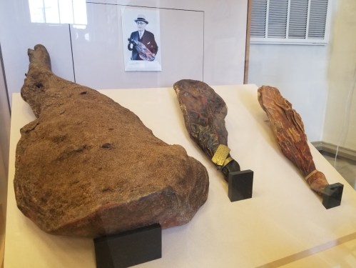Three large cured hams displayed in a glass case, with a black-and-white photo of a man holding a ham in the background.