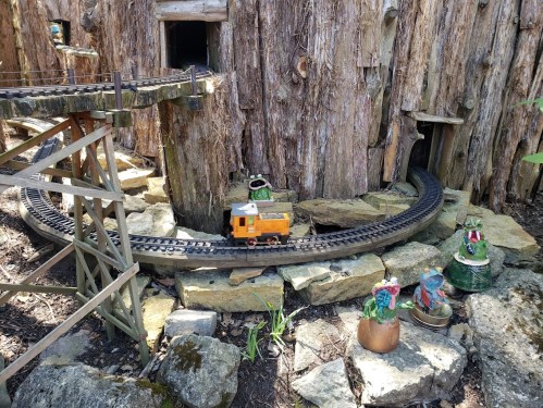 A miniature train on a winding track near a wooden structure, surrounded by rocks and small decorative figures.