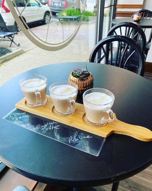 Three glass mugs of coffee on a wooden tray, displayed on a round table in a cozy café setting.