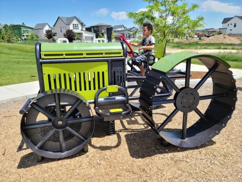 A child sits on a colorful playground tractor with large wheels, surrounded by grass and houses in the background.