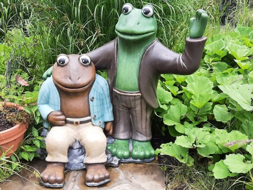 Two whimsical frog statues, one wearing a blue shirt and the other in a brown jacket, surrounded by greenery.