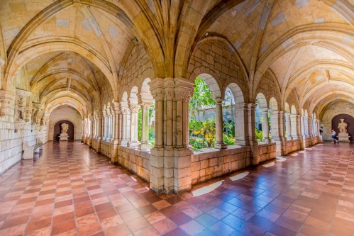 A serene stone corridor with arches, columns, and statues, leading to a lush garden in a historic building.