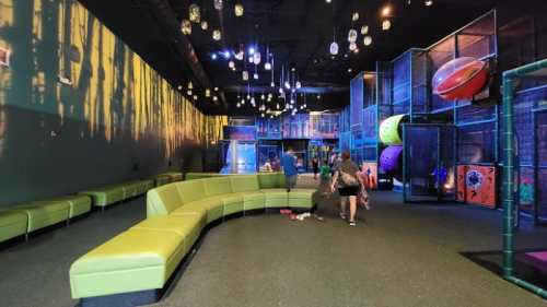 A colorful indoor play area with a green curved bench, climbing structures, and playful lighting. Children are playing.