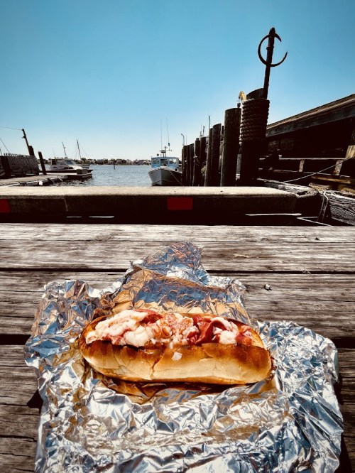 A lobster roll on foil sits on a wooden dock, with boats and a clear blue sky in the background.