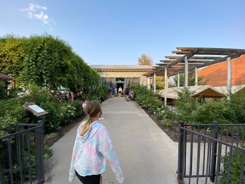 A girl walks down a path surrounded by greenery, leading to a building with people gathered outside.