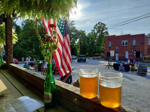 A sunny outdoor scene with two glasses of beer, flowers, and American flags, set in a lively gathering space.
