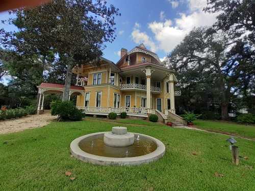 A large, historic yellow mansion with a fountain in the foreground, surrounded by lush greenery and trees.