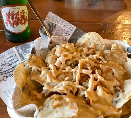 A plate of nachos topped with cheese and sauce, served with a bottle of Ale-8 soda on the side.
