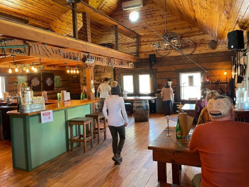 A rustic bar interior with wooden beams, tables, and patrons enjoying drinks in a cozy atmosphere.