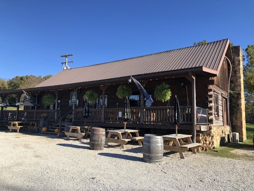A rustic wooden building with a metal roof, featuring outdoor seating and hanging plants, set against a clear blue sky.