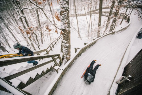 A child sleds down a snowy hill while another climbs stairs in a winter forest setting. Snow covers the ground and trees.