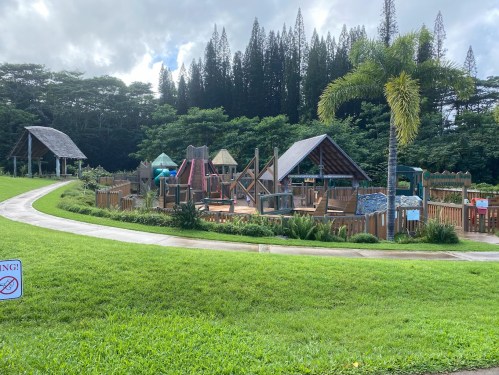 A colorful playground surrounded by greenery, featuring slides, climbing structures, and picnic areas under a blue sky.
