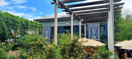 A vibrant outdoor café with lush greenery, colorful murals, and a pergola, set against a blue sky.