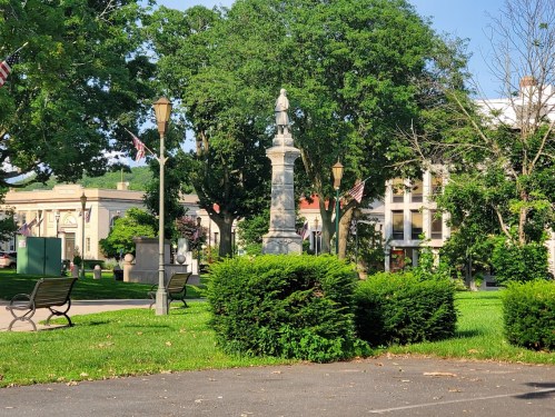 A park scene featuring a monument surrounded by trees, benches, and buildings, with American flags in the background.