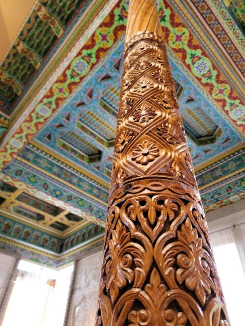 Intricately carved wooden pillar with vibrant, ornate ceiling patterns in the background.
