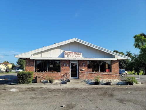 A brick building with a sign reading "Dixie Fried Chicken," surrounded by greenery and a clear blue sky.